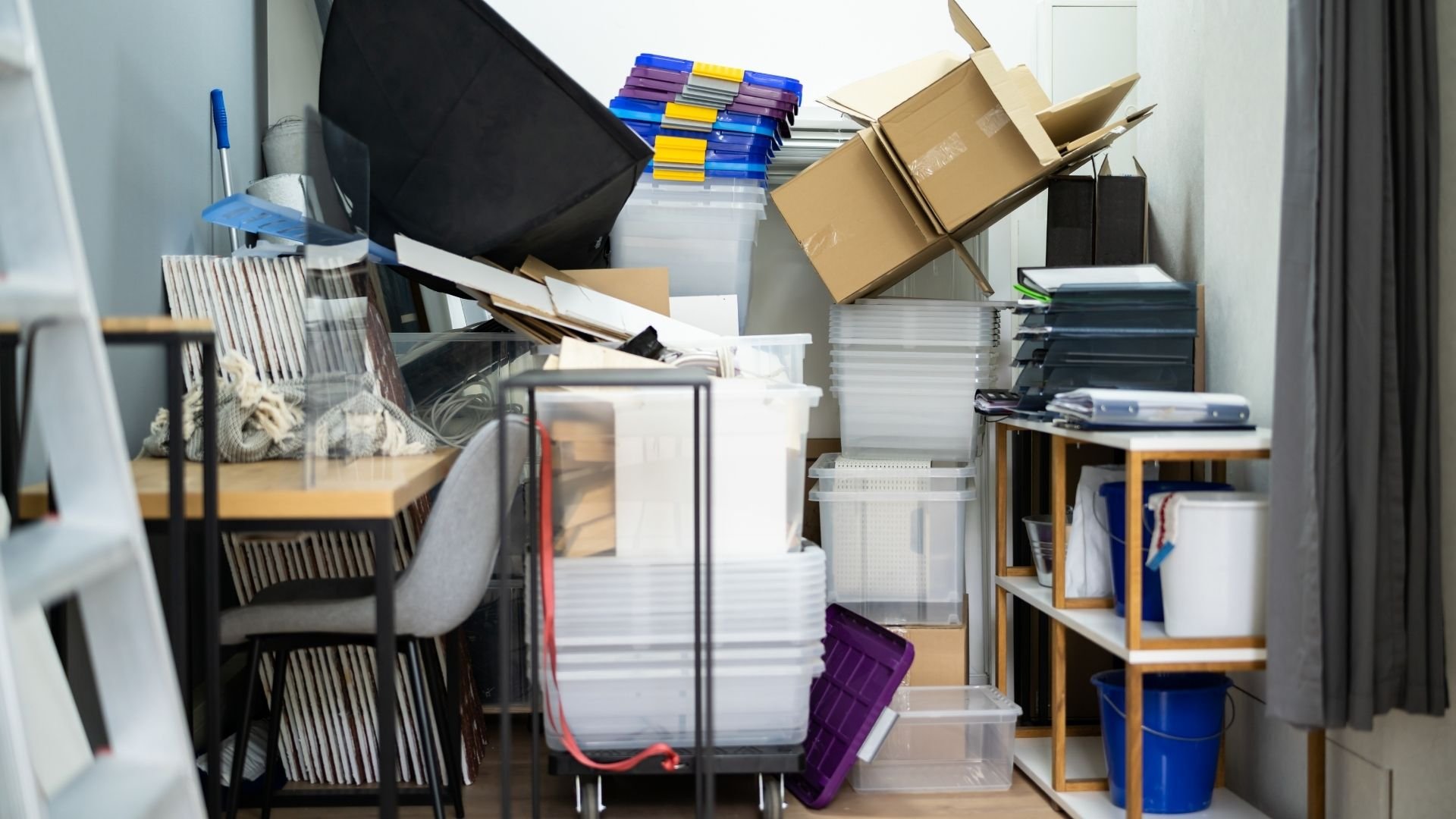 Cluttered storage room with stacked boxes, containers, office supplies, and disorganized shelving.