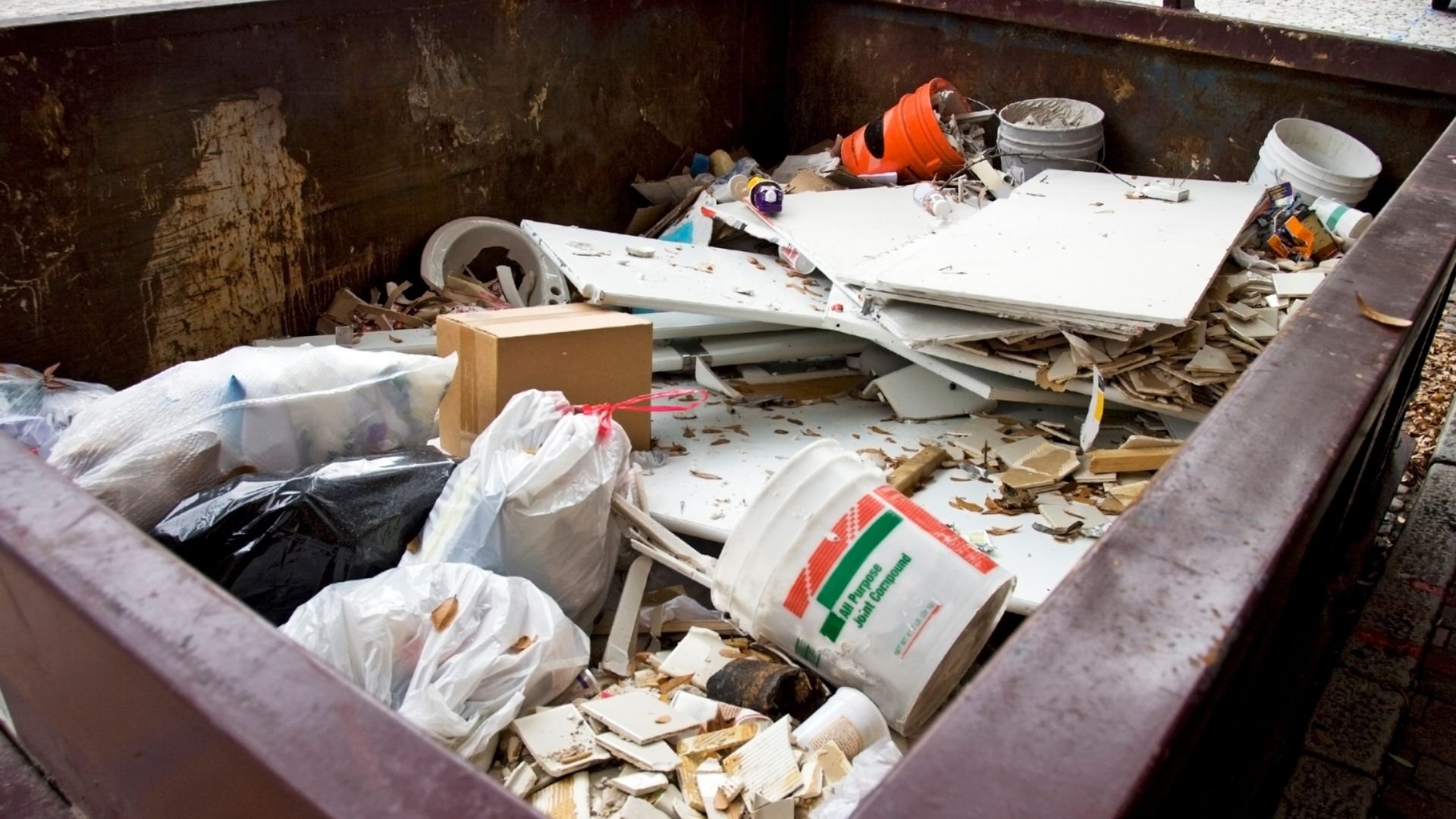 Dumpster filled with construction debris, trash bags, white buckets, wood scraps, and miscellaneous waste materials