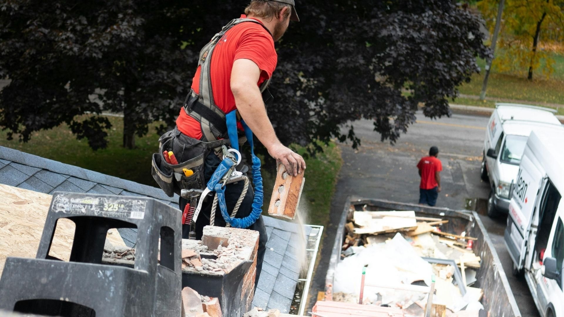Worker with safety harness and rope climbing on roof with debris and construction site below.
