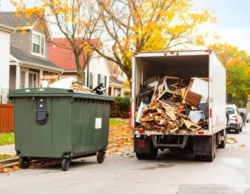 Junk removal truck loaded with wood debris and waste next to green dumpster on residential street.
