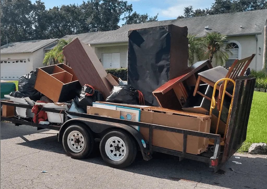 Junk removal trailer loaded with discarded furniture, boxes, and debris in driveway.
